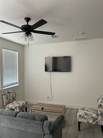 Lubbock apartment for rent - A living room with a grey couch, a chair with a floral pattern, a television on the wall, and a ceiling fan with lights.