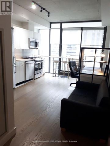 Ottawa condo for rent - A living room with a black couch and a kitchen area with white appliances.
