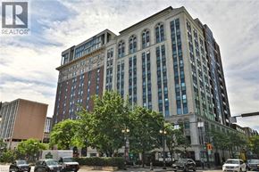 Hamilton condo for rent - A large, multi-story building with a mix of brick and glass windows is situated on a street corner with cars parked in front and a traffic light to the right.