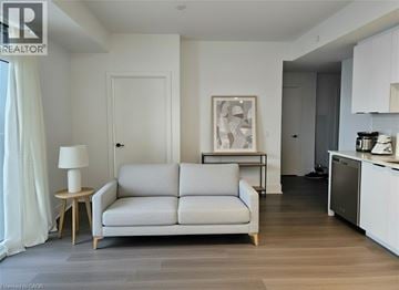 Kitchener condo for rent - A living room with a grey couch, a side table with a lamp, and a kitchen area with a fridge and stove.