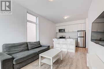 Oshawa (Samac) townhouse for rent - A grey couch sits in a white living room with a kitchen area in the background.