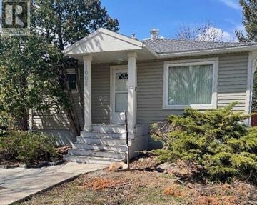 Lethbridge apartment for rent - A house with a grey siding and a white door is surrounded by greenery.