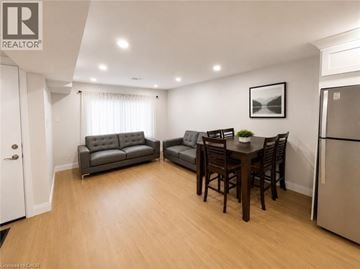 Kitchener apartment for rent - A well-lit living room with a grey sofa, a black leather armchair, a wooden dining table with chairs, and a refrigerator.