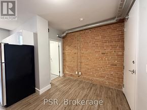 Toronto (Junction Area) condo for rent - A room with a brick wall and wooden flooring, with a black refrigerator on the left side.