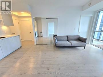 Toronto (Niagara) condo for rent - A spacious living room with a grey sofa and a kitchen area with white cabinets.
