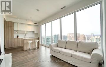 Toronto (Willowdale East) condo for rent - A spacious living room with a white sofa and a kitchen area in the background.