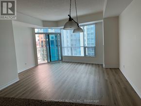 Barrie (City Centre) condo for rent - A large, empty room with wood flooring and a view of a city skyline through the windows.