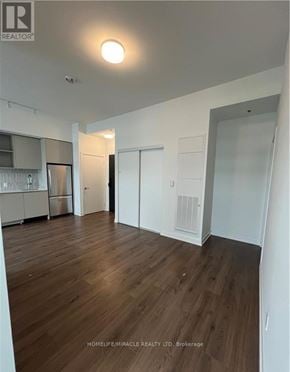 Oakville (Go Glenorchy) condo for rent - A room with wooden flooring and white walls, with a kitchen area visible in the background.