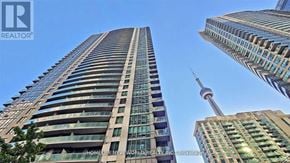 Toronto (Waterfront Communities) condo for rent - A tall building with a glass facade stands in front of a skyline with other buildings and a tower.