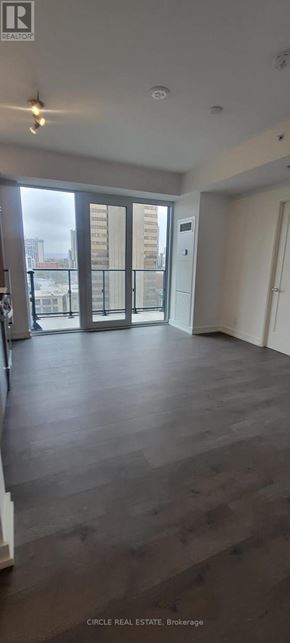 Hamilton (Corktown) condo for rent - A spacious room with wooden flooring and a balcony with a view of buildings outside.