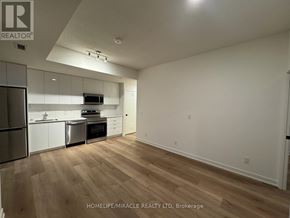 Brantford condo for rent - A well-lit, empty room with wooden flooring and a kitchen area to the left.