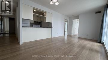 Toronto (Bendale) condo for rent - A spacious room with wooden flooring and white walls, with a kitchen area visible through a glass door.