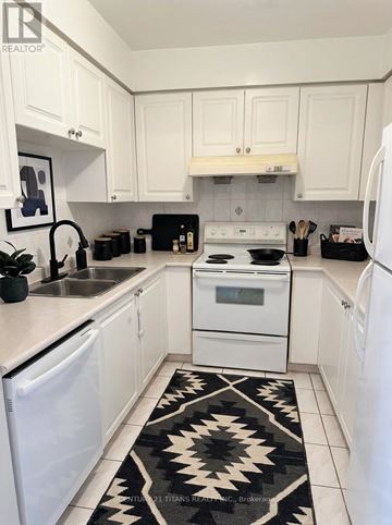 Toronto (Agincourt North) condo for rent - A kitchen with white cabinets and appliances, a black and white rug, and a framed picture on the wall.