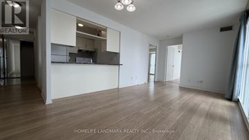 Toronto (Bendale) condo for rent - A spacious room with wooden flooring and white walls, with a kitchen area visible through a glass door.