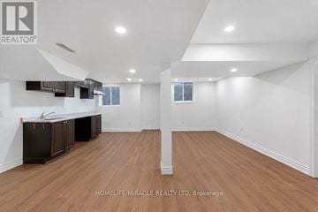 Oshawa (Taunton) apartment for rent - A spacious room with wooden flooring and white walls, featuring a kitchenette with dark cabinets and a countertop.