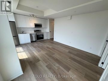 Brantford condo for rent - A spacious room with wooden flooring and white walls, featuring a kitchen area with a microwave and oven.