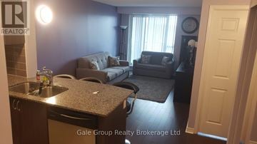 Brampton (Sandringham-Wellington) condo for rent - A living room with a grey couch and a kitchen area with a sink and a dishwasher.