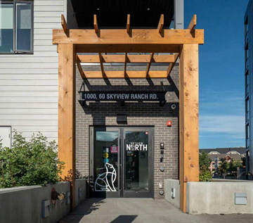 Calgary apartment for rent - The image shows the front of a building with a wooden structure above the entrance and a glass door with a bicycle symbol on it.