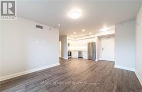 Niagara Falls (Hospital) condo for rent - A spacious room with wooden flooring and white walls, featuring a kitchenette in the background.