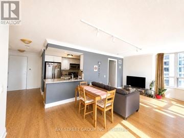 Toronto (Willowdale East) condo for rent - A living room with a dining table and chairs, a television, and a kitchen area in the background.