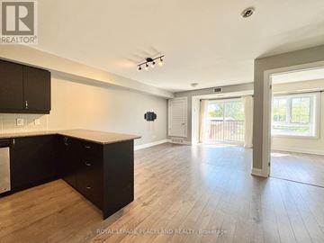 Whitchurch-Stouffville (Stouffville) condo for rent - A spacious room with wooden flooring and a kitchen area with black cabinets.