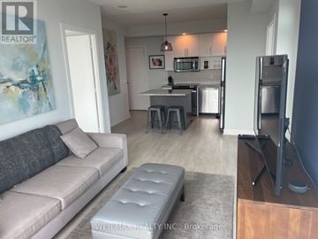Toronto (North St. James Town) condo for rent - A living room with a grey couch and a kitchen area in the background.