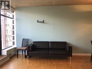 Vancouver apartment for rent - A black leather couch sits in a living room with a wooden floor and a wall mounted shelf.