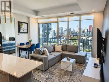 Toronto (South Riverdale) condo for rent - A living room with a grey couch and a large window overlooking a cityscape.