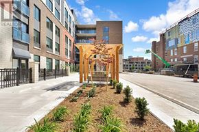 London East (East K) condo for rent - A wooden archway with leaves and flowers is in the middle of a landscaped area in front of a building.