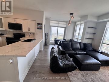 Mississauga (City Centre) condo for rent - A living room with a black leather couch and a kitchen area with white cabinets.