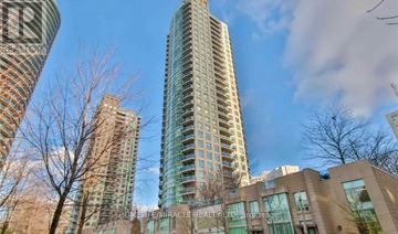 Mississauga (City Centre) condo for rent - A tall building with many windows stands in front of other buildings and trees.