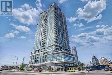 Mississauga (City Centre) condo for rent - A tall, modern glass building stands under a blue sky with scattered clouds.