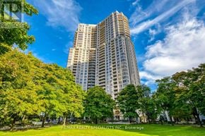 Mississauga (Hurontario) condo for rent - A tall building is surrounded by trees and has a blue sky in the background.