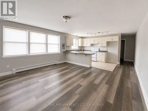 St. Catharines (E. Chester) multi-family for rent - A spacious room with wooden flooring and a kitchen area in the background.