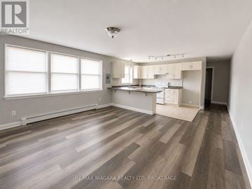 St. Catharines (E. Chester) multi-family for rent - A spacious room with wooden flooring and a kitchen area in the background.