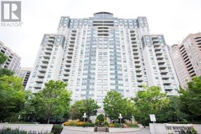Toronto (Willowdale East) condo for rent - A large building with a lot of windows and balconies is surrounded by trees and bushes.