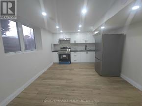 Brantford apartment for rent - A well-lit, empty room with wooden flooring and white walls, featuring a kitchenette with a refrigerator, sink, and cabinets.