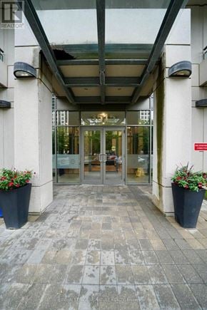 Mississauga (Fairview) condo for rent - The entrance to a building with glass doors and two large planters with red flowers on either side.