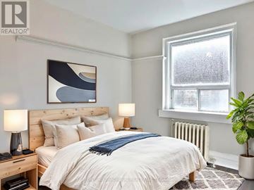 Toronto (Little Portugal) condo for rent - A bedroom with a large bed, a nightstand with a lamp, a potted plant, and a window with a view of a snowy landscape.