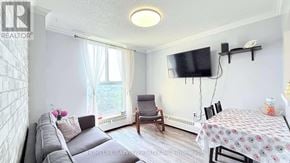 Toronto (Black Creek) condo for rent - A living room with a grey sofa, a small table with a floral tablecloth, and a television mounted on the wall.