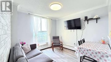 Toronto (Black Creek) condo for rent - A living room with a grey sofa, a small table with a floral tablecloth, and a television mounted on the wall.