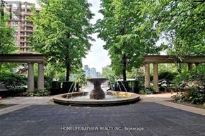 Toronto (Willowdale East) condo for rent - A fountain in the middle of a park with trees and buildings in the background.
