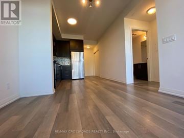 Markham (Wismer) condo for rent - A spacious room with wooden flooring and a kitchen area in the background.