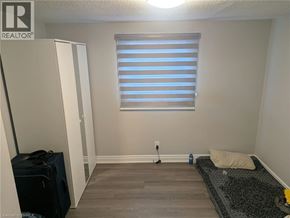 Brantford apartment for rent - A room with a window covered by horizontal blinds, a rug on the floor, and a suitcase on the left side.