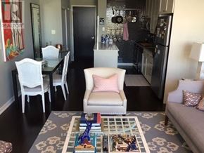 Toronto (Niagara) condo for rent - A living room with a grey couch, a coffee table with a book and a blue figurine on it, a white chair, a dining table with four chairs, a kitchen area with a refrigerator, a sink, and