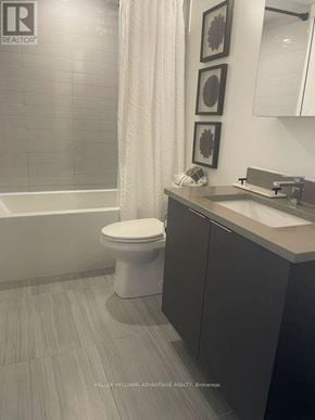 Toronto (Agincourt South-Malvern West) condo for rent - A white toilet sits in a bathroom with a grey tile wall and a sink with a mirror above it.