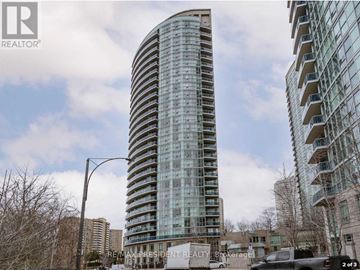 Mississauga (City Centre) condo for rent - A tall glass building with a curved design stands in front of a cloudy sky.