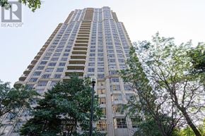 Mississauga (Hurontario) condo for rent - A tall building with many windows is surrounded by trees.
