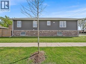 Tecumseh apartment for rent - A small tree stands in a grassy area in front of a grey house.