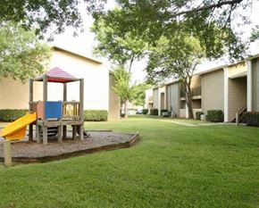 Hurst apartment for rent - A playground with a slide and a wooden structure is surrounded by a grassy area and apartment buildings.